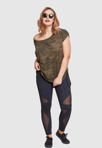 Olivenfarget kamuflasje oversized topp, svarte leggings med meshutskjæringer, og svarte sneakers. Solbriller fullfører den sporty stilen.