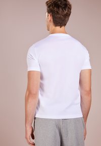 Biała koszulka z krótkim rękawem w miękkim materiale, z okrągłym dekoltem, noszona z szarymi spodniami. Widok z tyłu pokazuje gładką fakturę i dopasowany fason.