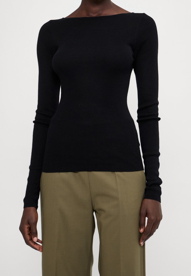Personne portant un haut noir ajusté à manches longues et un pantalon ample de couleur vert olive, debout devant un fond uni clair.