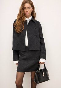 Mörkgrå ulljacka med en kortad design och dubbelknappning, ihopparad med en matchande kjol. Håller en svart handväska med silverdetaljer.