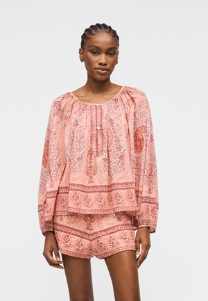 Mujer vistiendo una blusa suelta de manga larga con estampado de paisley rosa y pantalones cortos a juego, posando frente a un fondo claro y neutro.