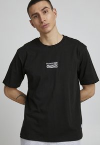 Musta puuvillapaita lyhyillä hihoilla, jossa on valkoinen brodeerattu merkki "SLD-EST.1999" ja raidallinen yksityiskohta rinnassa.