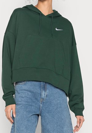 Osoba w ciemnozielonej bluzie z kapturem Nike z troczkami i jasnoniebieskich dżinsach, ręce swobodnie ułożone wzdłuż ciała.