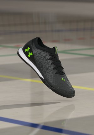 Chaussure de sport d’intérieur noire avec logos Under Armour vert néon, dessus à motifs et semelle blanche, flottant au-dessus d’un sol de gym avec des lignes de terrain.