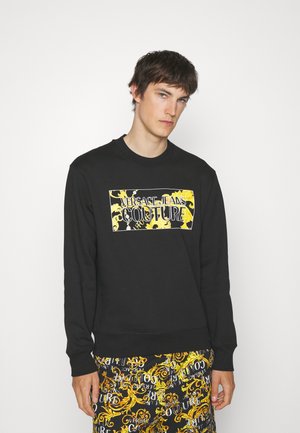 Felpa nera con una grafica rettangolare gialla e nera che riporta la scritta "Versace Jeans Couture". Collo e polsini a coste, vestibilità comoda.