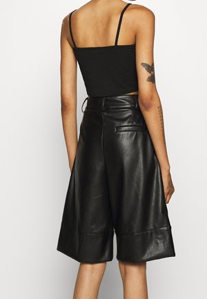 Rückansicht einer Person, die ein schwarzes Spaghettiträgertop und hoch taillierte schwarze Culotte-Shorts aus Leder trägt, mit Tätowierungen am rechten Arm.