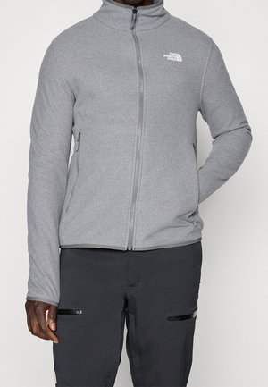 Person trägt eine hellgraue Fleecejacke mit durchgehendem Reißverschluss, zwei Reißverschlusstaschen und dunkle Hosen mit horizontalen Reißverschlusstaschen.