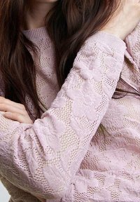 Lichtroze gebreide trui met bloemmotieven, gestructureerde mouwen en een geribde boord. De stof heeft een ademend, open geweven ontwerp.