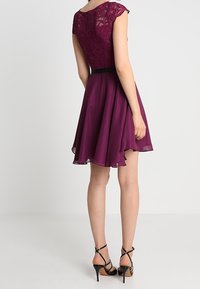 Femme portant une robe violette sans manches, longueur genoux, avec un corsage en dentelle et une jupe fluide, associée à des sandales à talons hauts noires à lanières, debout.