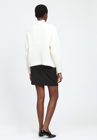 Weißer, strukturierter, kurzer Pullover kombiniert mit einem schwarzen, figurbetonten Rock. Die Schuhe sind glänzend schwarz mit Schnallen und präsentieren ein elegantes Design.