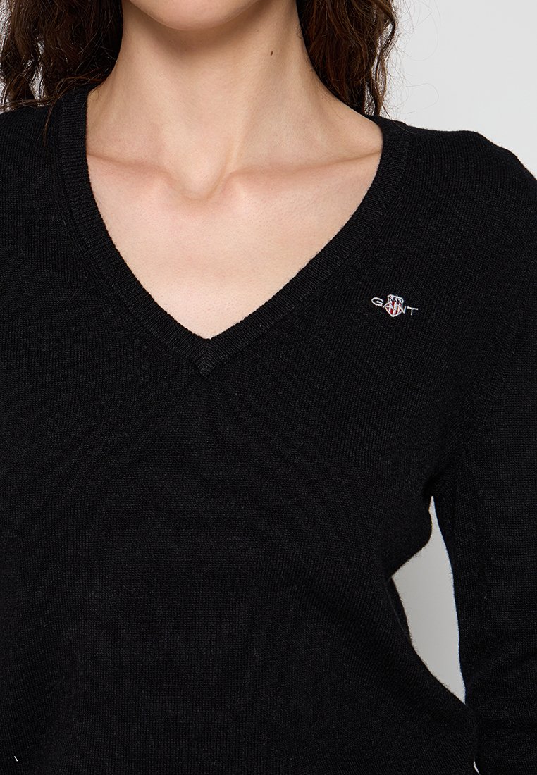 Schwarzer V-Ausschnitt-Pullover aus weichem Material, mit einem kleinen gestickten Logo in Rot und Weiß auf der linken Brust. Strukturierter Strickstoff.