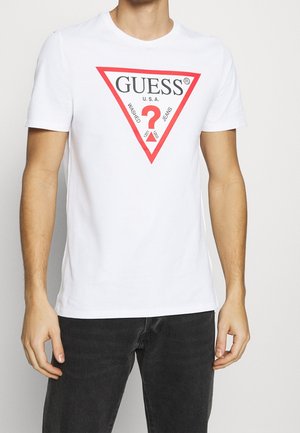 Homme portant un t-shirt blanc à manches courtes avec un triangle rouge et le texte noir "GUESS U.S.A." imprimé sur le devant, associé à un jean foncé.