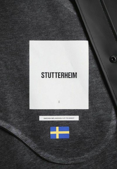 Grijze stof met een gladde textuur. Labels omvatten "STUTTERHEIM," maat "S," en de Zweedse vlag. Zwarte accent langs de rand.