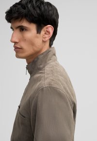 Taupe half-zip jas van zachte stof met een gladde, textuurlijke afwerking. Voorzien van een ritsborstzak en een hoge kraag, slim fit.