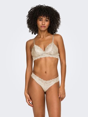 Woman with curly hair wearing beige lace lingerie set, standing against a plain light background, facing forward with relaxed posture.