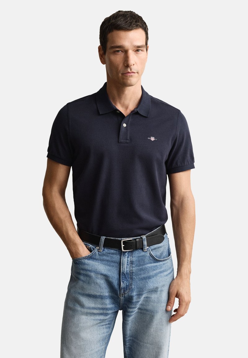 Marineblå pikéskjorte med korte ermer, to-knappers stolpe, brodert logo på brystet, kombinert med lyseblå denimjeans og svart belte.