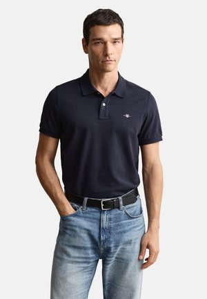 Polo bleu marine, manches courtes, patte de boutonnage à deux boutons, logo brodé sur la poitrine, porté avec un jean en denim bleu clair et une ceinture noire.