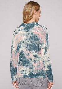 Pullover a maglia rigata in teal con motivo astratto rosa e bianco, con colletto arrotondato e maniche lunghe, abbinato a jeans grigi.