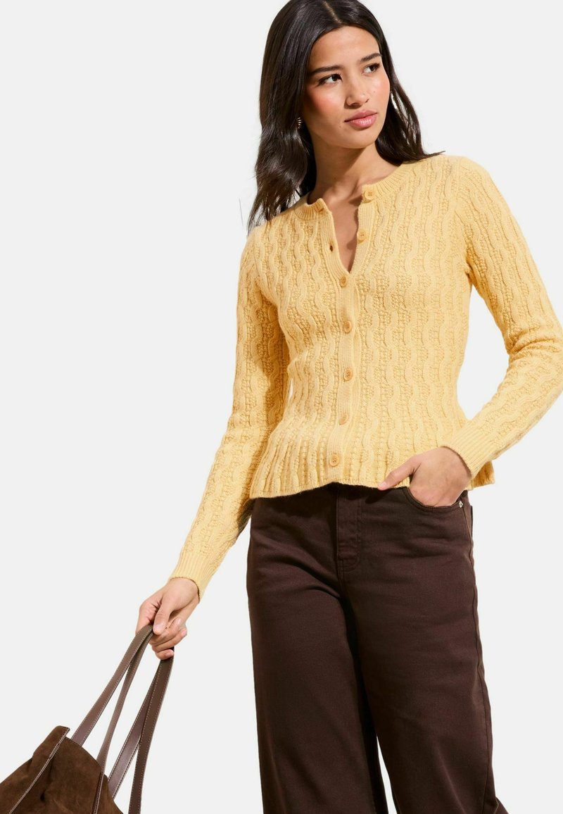 Mujer vistiendo un cárdigan amarillo con textura y botones, y pantalones marrón oscuro, sosteniendo un bolso tote de ante marrón en una mano.