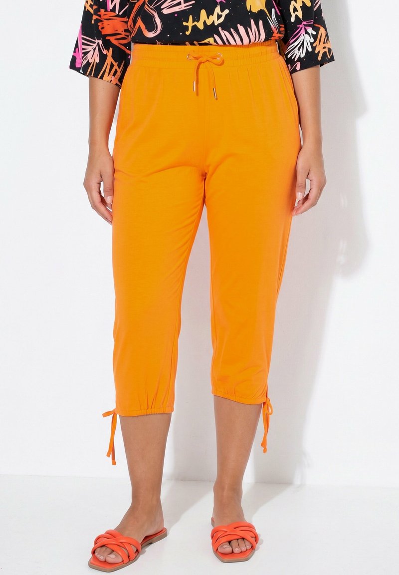 Femme portant un pantalon court orange vif avec taille à cordon et liens à la cheville, associé à des sandales orange et un haut à motifs.