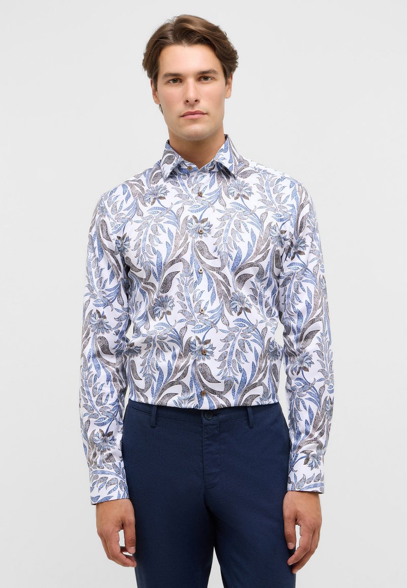 Witte blouse met een blauw en bruin bloemenpatroon, flexibele stof, lange mouwen en een button-down kraag. Gemaakt met marineblauwe broek.