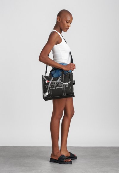 Svart kanvas totebag med hvite sømmer og grafisk tekst. Har korte håndtak og avtagbar skulderrem. Teksturert finish.