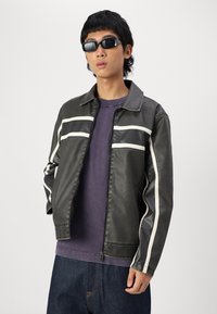 Black faux leather jacket with white accent stripes, a collared neck, and zip closure. Worn over a purple shirt, with visible blue denim pants.