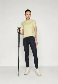 Κίτρινο μπλουζάκι κοντομάνικο και Ναυτικές κολάν με τσέπες, σε συνδυασμό με μπεζ παπούτσια. Το άτομο κρατά ένα μαύρο περιπατητικό μπαστούνι. Απλό φόντο.