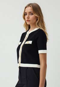 Cardigan nero a maniche corte con accenti bianchi a costine, caratterizzato da due tasche sul petto e bottoni dorati lungo la parte anteriore.