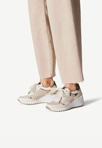 Sneaker bianchi e dorati con un design a materiali misti, caratterizzati da tessuto, accenti in pelle e motivi intricati, mostrati con pantaloni aderenti.