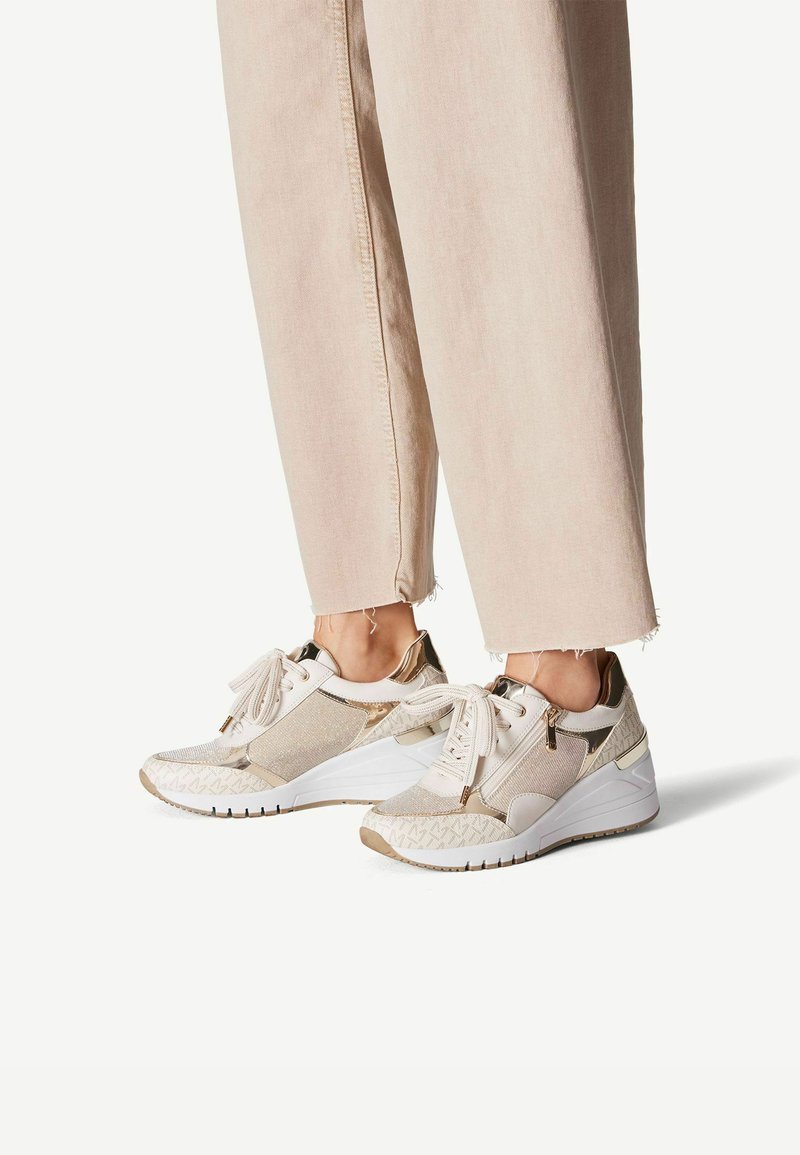 Sneaker bianchi e dorati con un design a materiali misti, caratterizzati da tessuto, accenti in pelle e motivi intricati, mostrati con pantaloni aderenti.