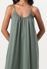 Sage zelené šaty bez rukávů s volným střihem, staženým výstřihem a měkkou tkaninou. Obsahují tenké ramínka a boční kapsy.