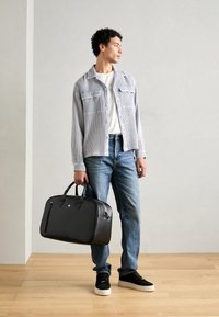 Svart lær duffelbag med glatt tekstur og dobbelt håndtak, parret med en grå rutete jakke, hvit t-skjorte og blå jeans.