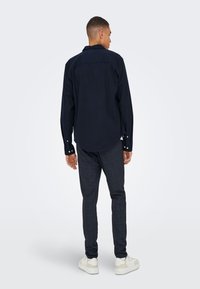 Temno modra srajca z okroglim ovratnikom, dolgimi rokavi in srebrnimi gumbi; kombinirana z sivimi karirastimi hlačami in belimi supergami.