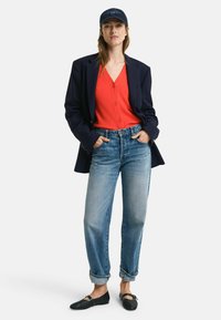 Navy blazer over a red cardigan, light blue relaxed-fit jeans, and black woven flats. Model wears a dark cap. Simple background.