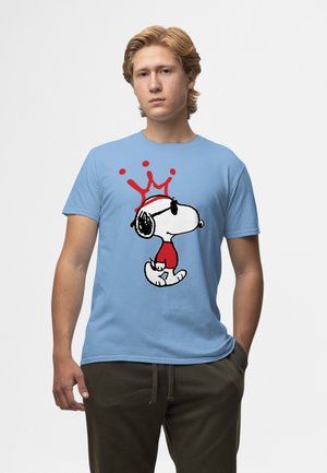 Jonge man die een lichtblauw T-shirt draagt met een Snoopy-afbeelding in rood en zwart, en een rood kroondesign boven Snoopys hoofd.