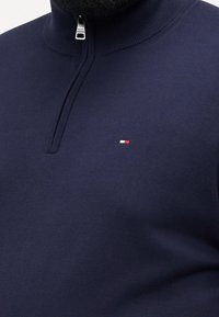 Pull en maille bleu marine à fermeture éclair partielle argentée avec petit logo rectangulaire rouge, blanc et bleu sur le côté gauche de la poitrine.