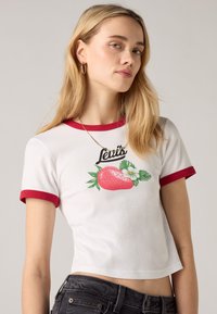 Valkoinen lyhythihainen t-paita punaisilla pääntie- ja hihasaumakujilla, jossa on painettu mansikka- ja kukkakuosi sekä "Levi's" -teksti graafisen kuvan yläpuolella.