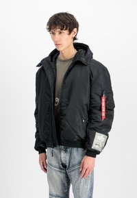 Melnā pufotā bomber jakā ar kapuci, priekšējo rāvējslēdzēju, ribotām piedurknēm un sarkano birku uz piedurknes. Tai ir kabata ar grafisku detaļu.