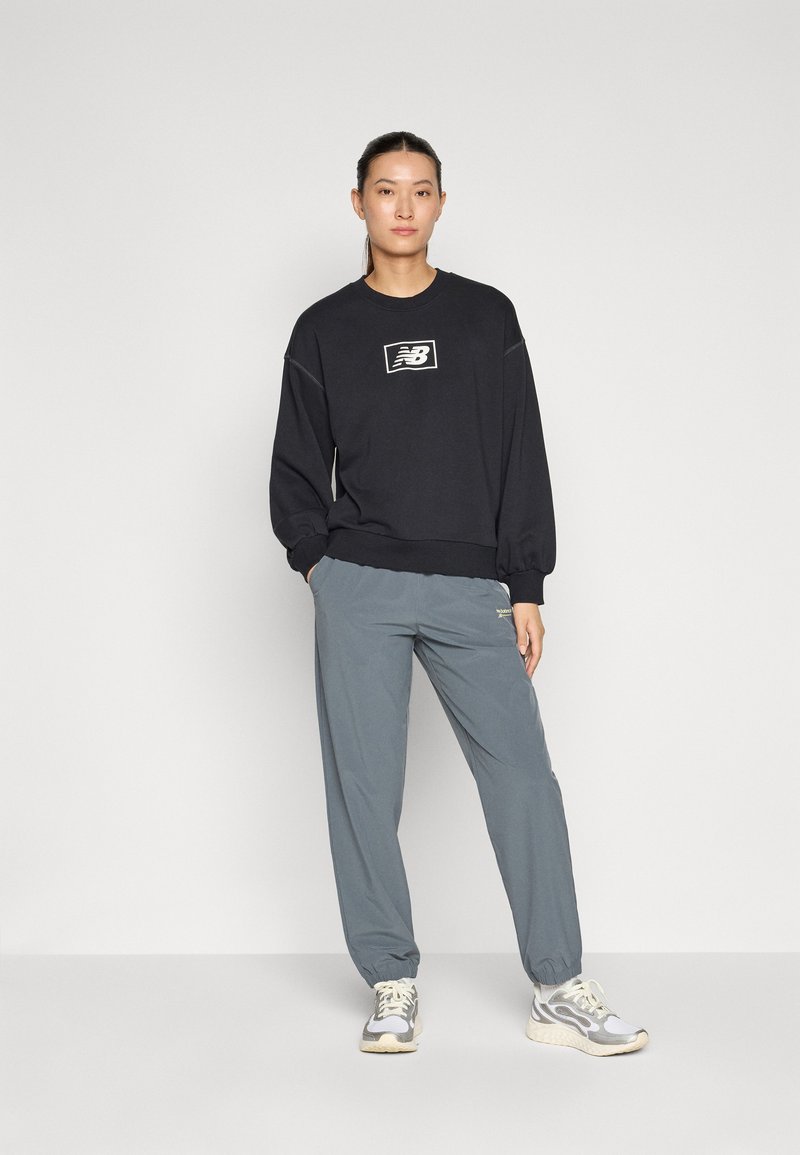 Felpa nera con logo bianco di New Balance, abbinata a pantaloni della tuta grigi chiari con polsini elastici, completati da sneakers grigie.