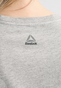 Camiseta de algodón gris con cuello acanalado, que presenta un logo de Reebok impreso en negro y un acento triangular en la parte posterior.