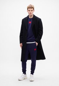 Granatowy sweter z czerwoną naszywką, w połączeniu z dopasowanymi dresami i długim czarnym płaszczem, uzupełniony białymi sneakersami.