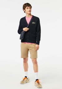 Marinblå ribbad cardigan med knappar, buren över en rosa T-shirt, kombinerad med beige shorts och flerdelade sneakers. Svart axelremsväska.