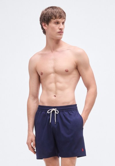 Marineblå badebukser med elastisk midjebånd og snor, med en liten rød logo, laget av glatt, lett stoff.