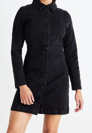 Kvinde iført en tætsiddende sort denimkjole med knapper, lange ærmer og krave, stående foran en ensartet hvid baggrund.