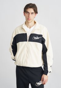 Αντιανεμικό μπουφάν Nike σε εκρού και μαύρο, με φερμουάρ, διαθέτει το λογότυπο σε λευκό. Κομμένο σε άνετη γραμμή με ελαστικές μανσέτες και τσέπες.