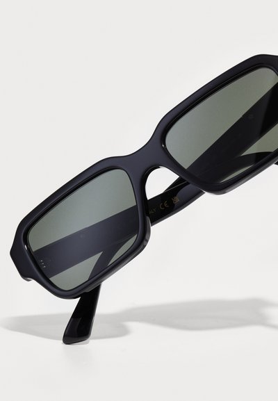 Lunettes de soleil noires surdimensionnées avec une large monture en plastique et des verres teintés foncés. Elles présentent des branches droites et un logo de marque subtil sur la face interne.