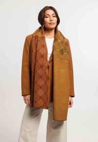 Écharpe à motifs marron et jaune en matériau tissé texturé, drapée sur les épaules d'un modèle, avec des designs géométriques et une finition douce.