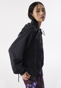 Chaqueta bomber negra con un acabado texturizado, puños elásticos, cordón en el cuello y un corte relajado. Combinada con pantalones morados estampados.