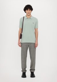 Világos zöld rövidujjú poloing, kis logóval, szürke kockás nadrággal és fekete cipővel párosítva, valamint egy barna crossbody táskával.